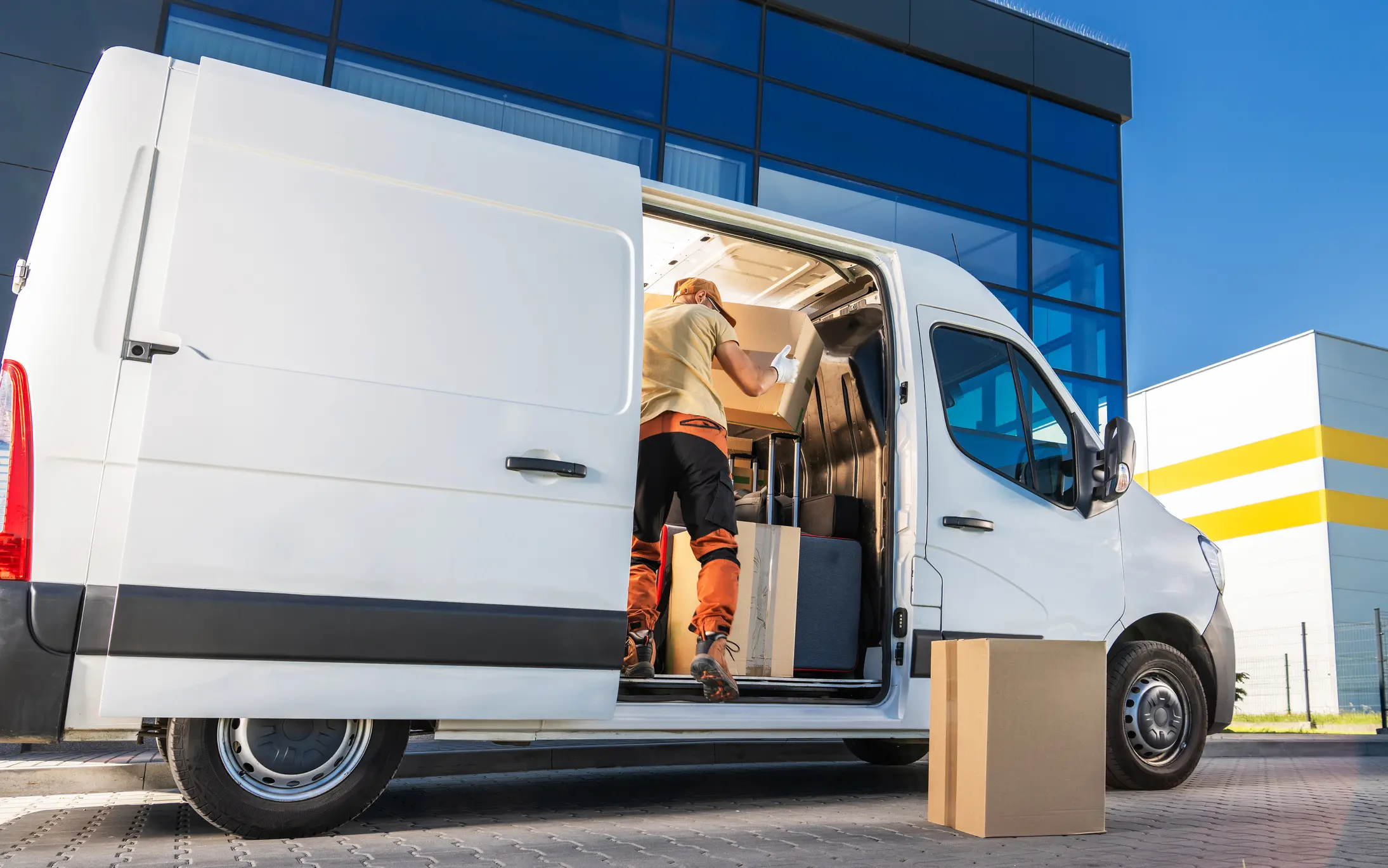 A delivery worker in an orange shirt and pants is loading boxes into a white van. The van is parked in front of a building with blue and white glass panels.